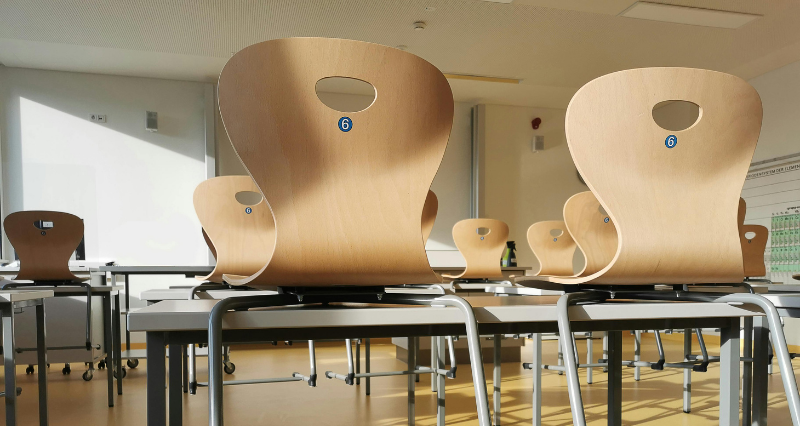 A photograph of an empty classroom with chairs on desks. Warm summer light fills the room.